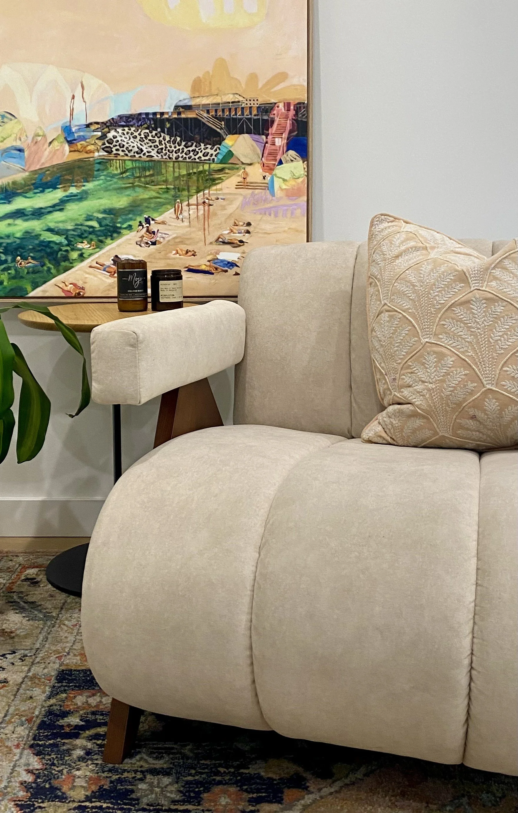 Close-up of a light beige armchair with a textured decorative pillow in a living room, with a large colorful beach-themed painting hanging on the wall behind it, and a small side table with two containers on it.