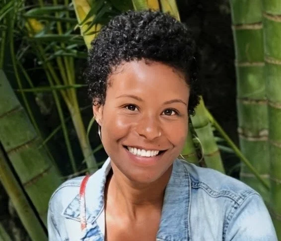 Smiling woman with of color, African, short curly hair wearing a denim jacket, standing in front of green bamboo plants.