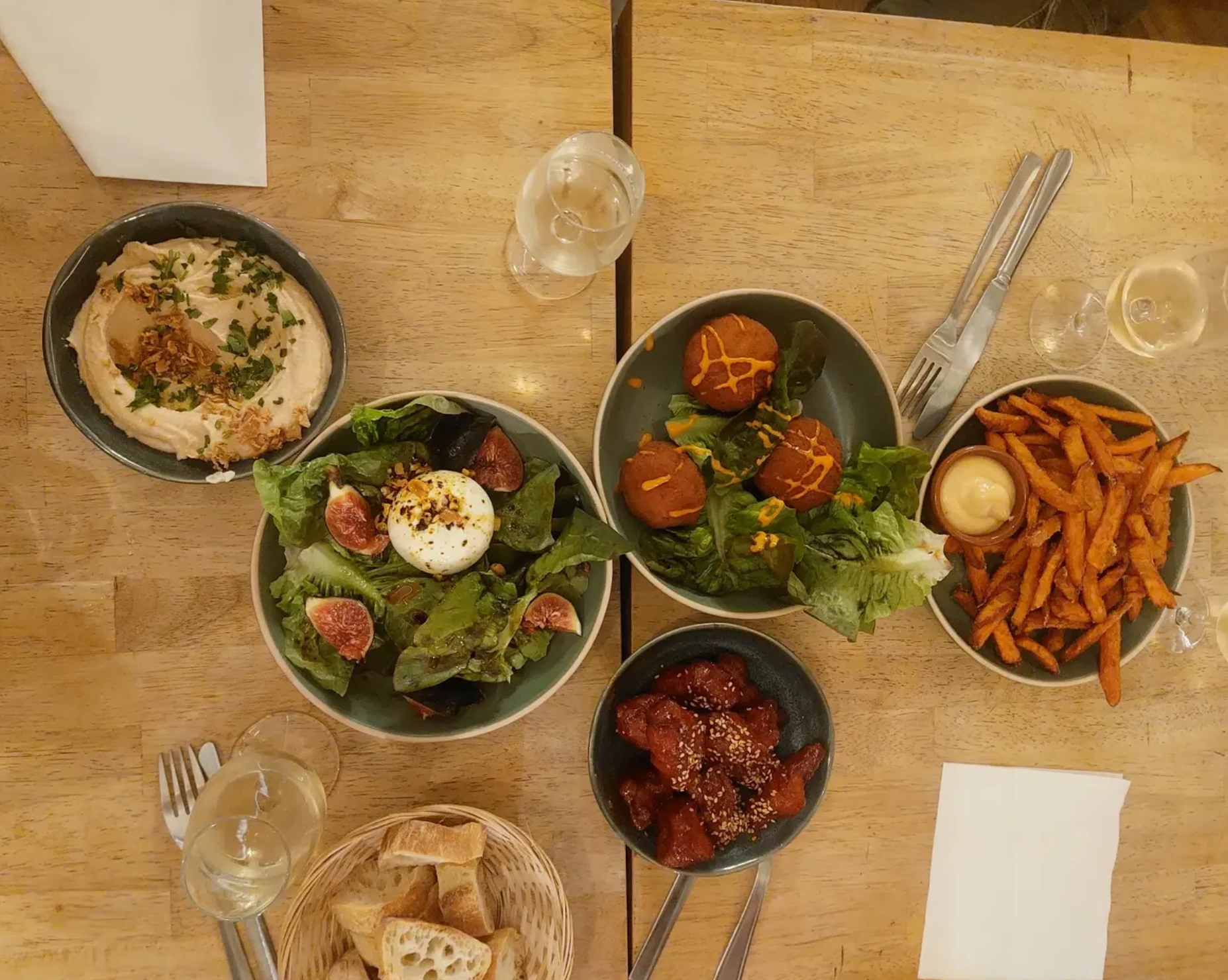 Un repas composé de plusieurs plats sur une table en bois. Deux salades, une avec œuf dur, figues, et légumes verts, l'autre avec des boulettes de falafel, une portion de frites, un bol de poulet épicé, un plat de porc ou poulet en sauce, un panier de pain, et deux verres de vin blanc.