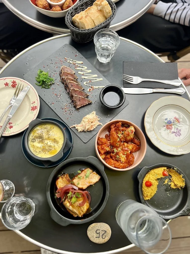 Une table avec un assortiment de plats et boissons, comprenant du steak tartare, des noix de pochtron, une soupe, un plat de fruits de mer épicés, un plat de poisson garni de légumes, et plusieurs assiettes avec des pains, une bouteille d'eau et un verre de vin.