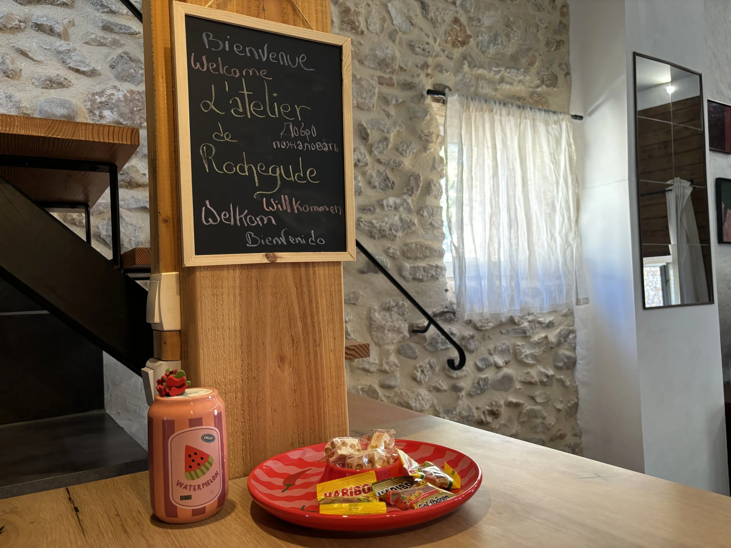 Un tableau noir avec un cadre en bois indique en français et en anglais l'accueil à l'atelier, avec un fond en pierre. Sur la table en bois au premier plan, il y a une bouteille de jus de watermelon, un plateau rouge avec des bonbons, et des sucettes.