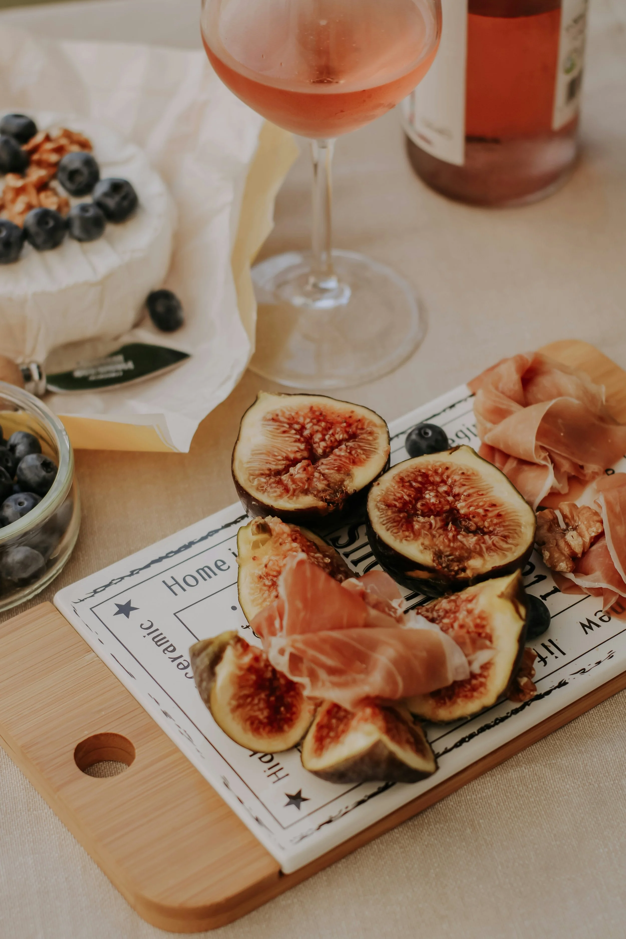 Une planche à découper en bois avec des figues coupées, du jambon cru, et une assiette avec des myrtilles, une tasse en verre avec du rosé, et un pot de confiture en arrière-plan.