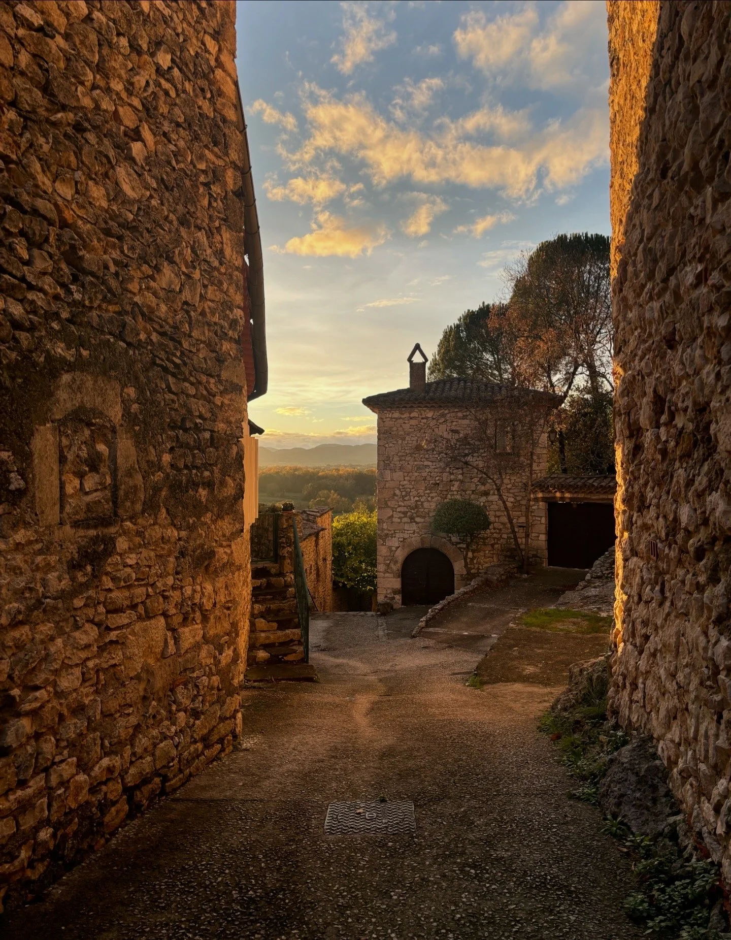Une ruelle pavée bordée de murs en pierre, avec une vieille maison en pierre au fond, sous un ciel orange au coucher du soleil.