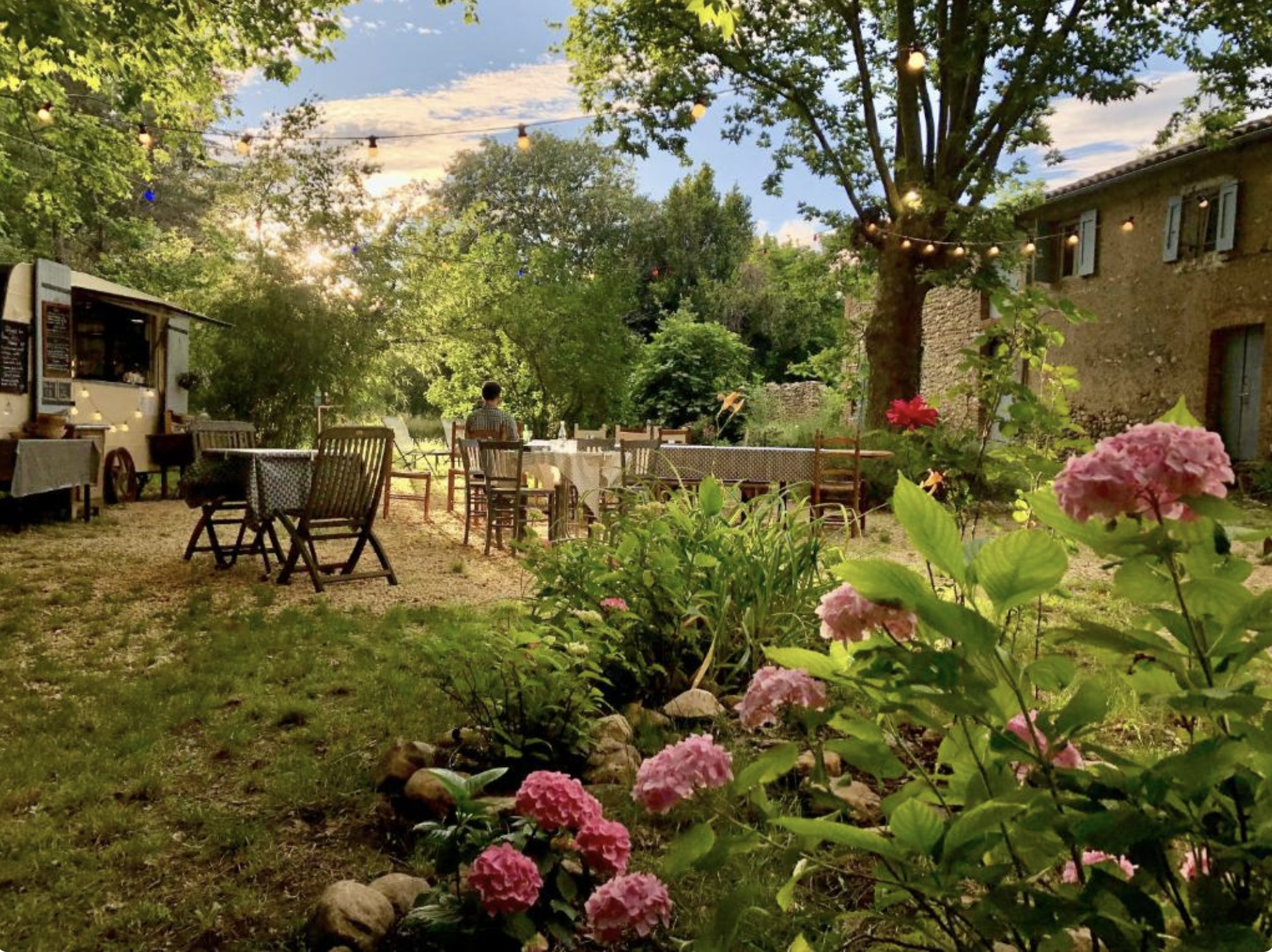 Une terrasse extérieure avec plusieurs tables et chaises sous un grand arbre. Des guirlandes lumineuses sont suspendues entre les arbres. En arrière-plan, une maison en pierre avec des volets bleus, entourée de verdure et de fleurs. Le soleil commence à se coucher, créant une ambiance chaleureuse et paisible.