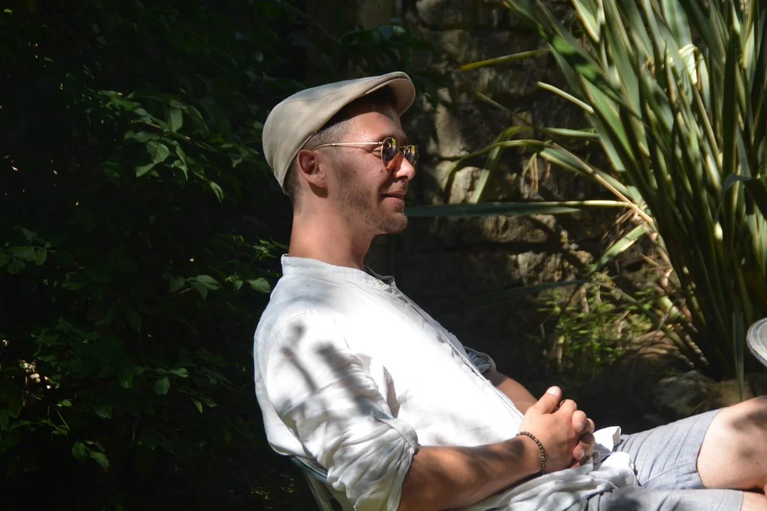 Un homme assis dans un extérieur ensoleillé, portant une chemise blanche, un chapeau beige, des lunettes de soleil, et un bracelet, dans un environnement verdoyant avec des plantes et des rochers.
