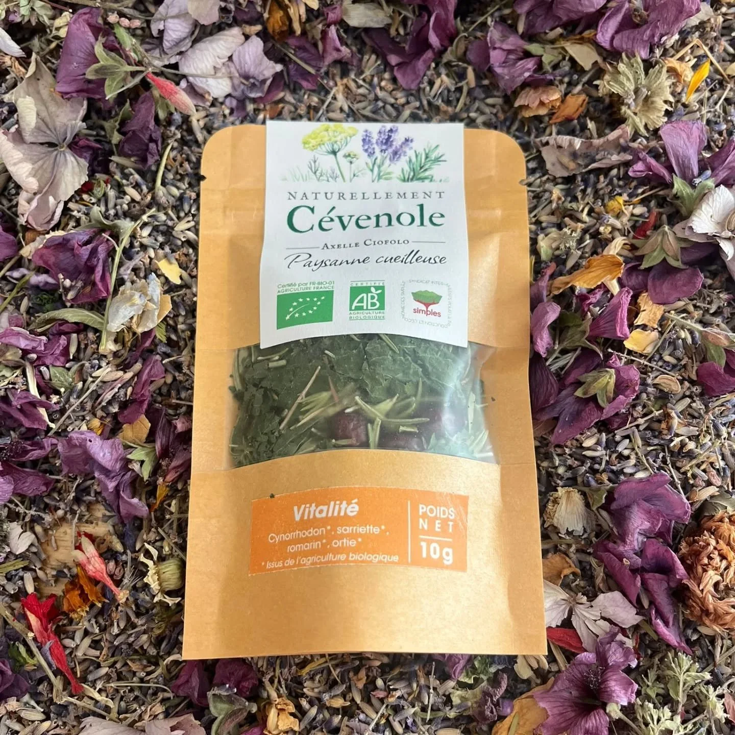 Paquet d'herbes aromatiques biologiques Cévenol avec étiquettes d'agriculture biologique française, Vegan, 10g, posé sur un fond de fleurs séchées violettes, roses, oranges et autres couleurs.