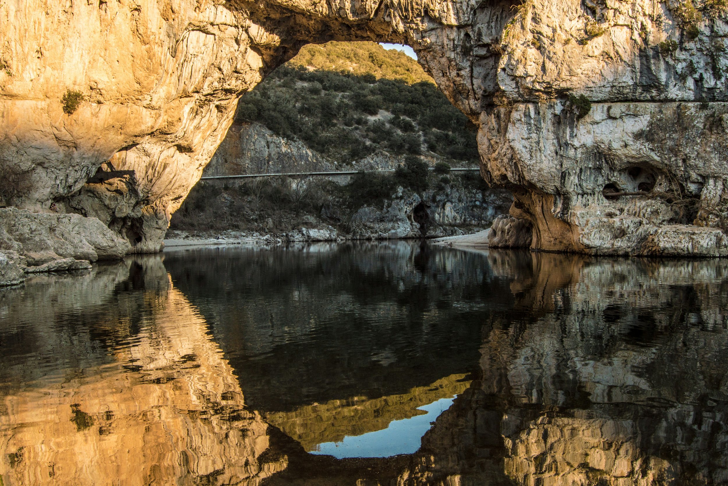 Vaste formation rocheuse avec une arche naturelle au-dessus d'une rivière, entourée de végétation et reflétée dans l'eau.