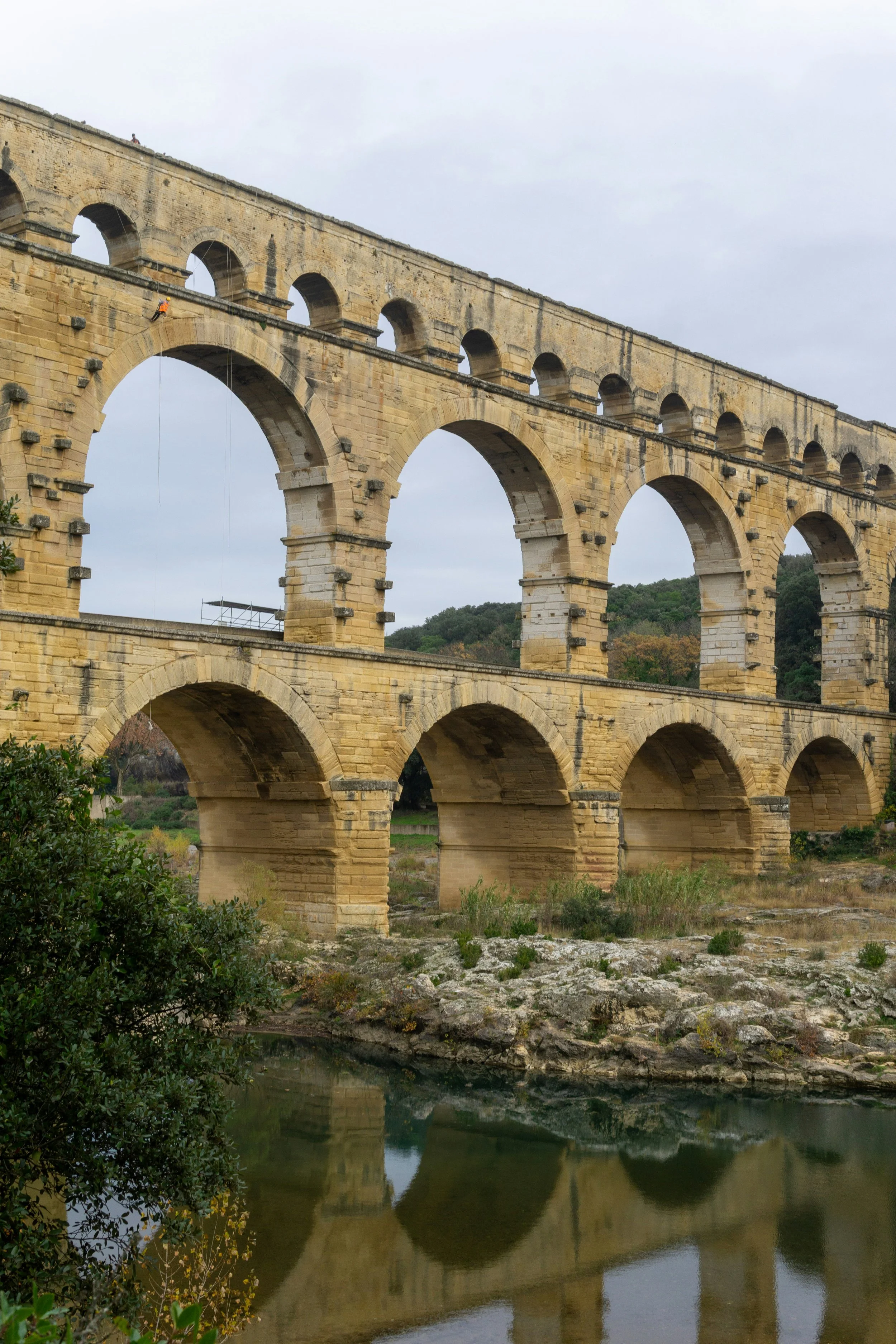 Un aqueduc en pierre avec plusieurs arches, reflété dans l'eau en dessous, avec un ciel nuageux et des collines en arrière-plan.