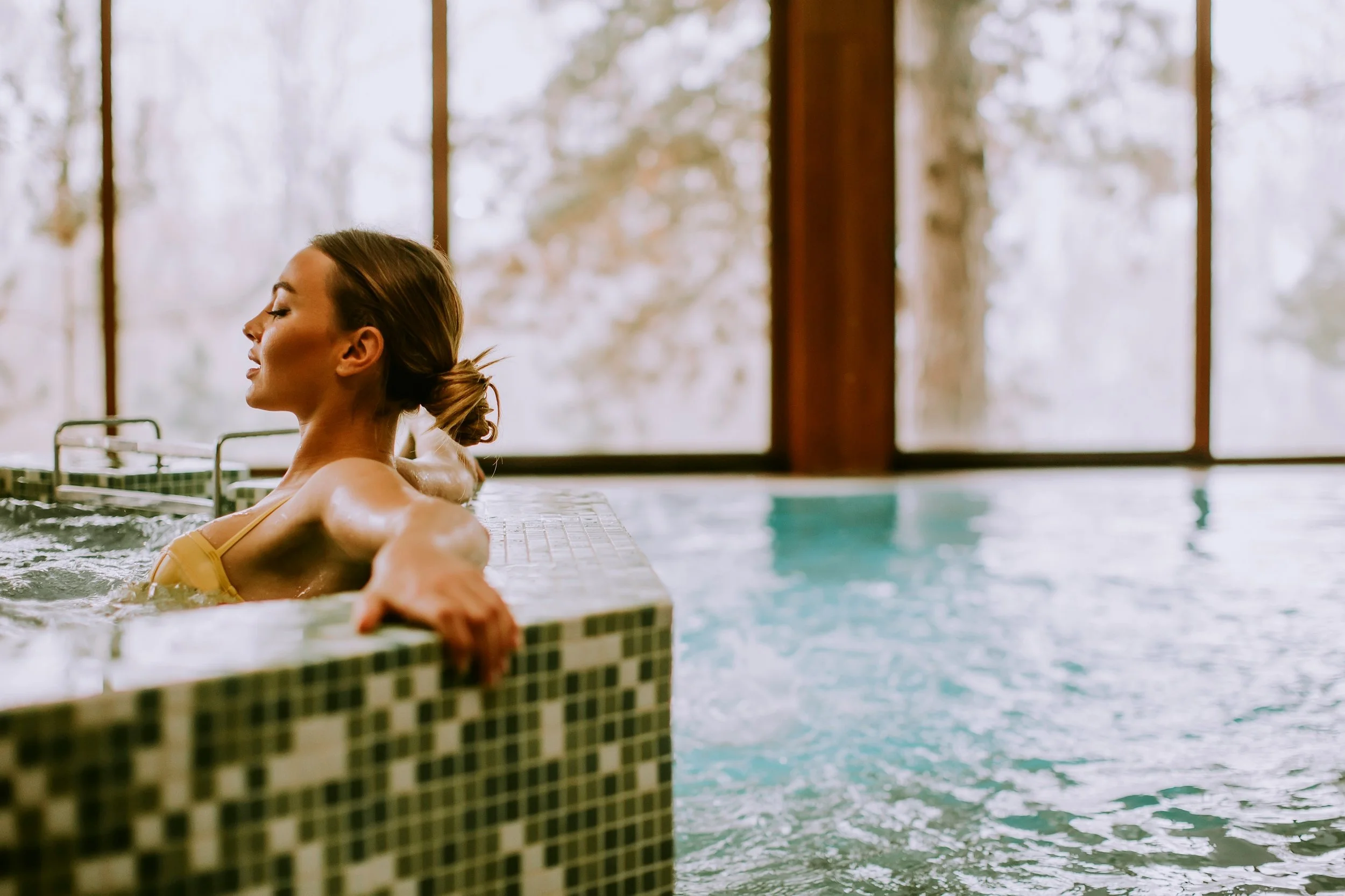 Femme détendue dans une baignoire en train de profiter d'un moment de relaxation, avec une vue sur des arbres à travers de grandes fenêtres en arrière-plan.