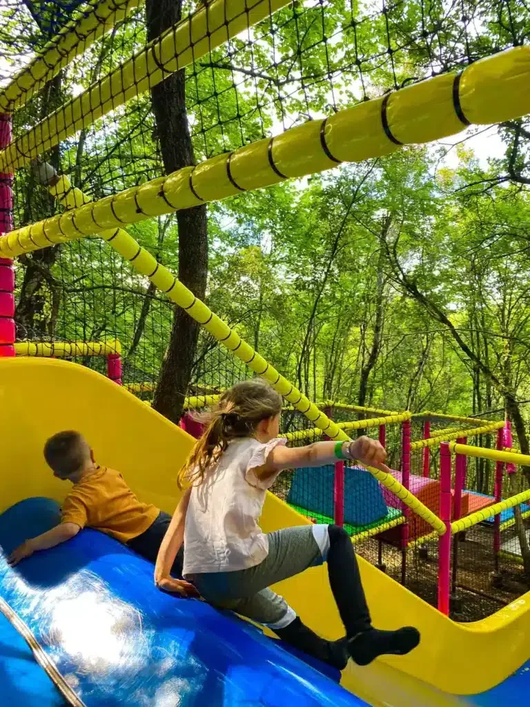 Deux enfants jouent sur une glissade colorée en bois avec un cadre de filet pour l'escalade dans un parc forestier.
