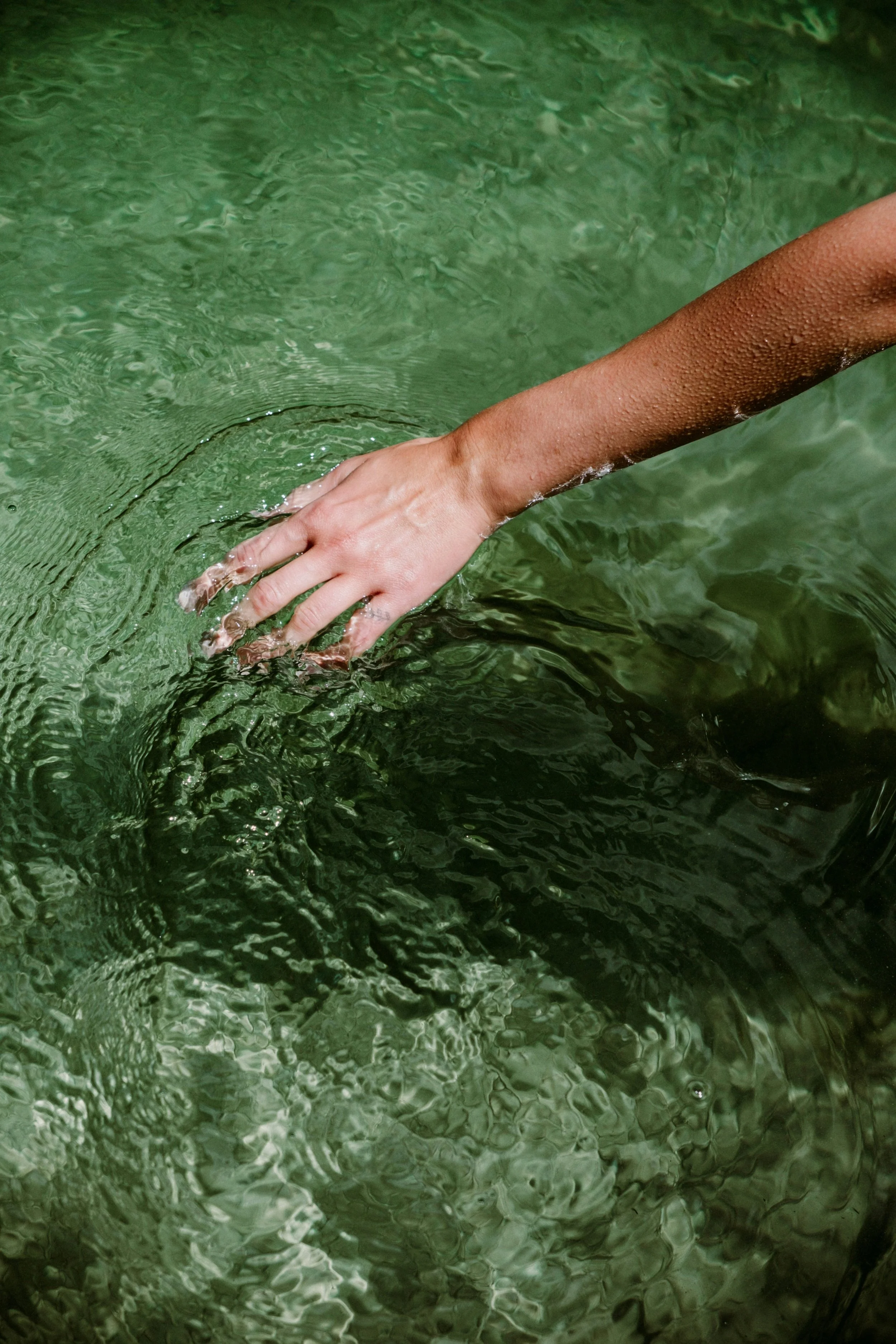 Une main touche la surface d'une eau verte, créant des ondes circulaires.