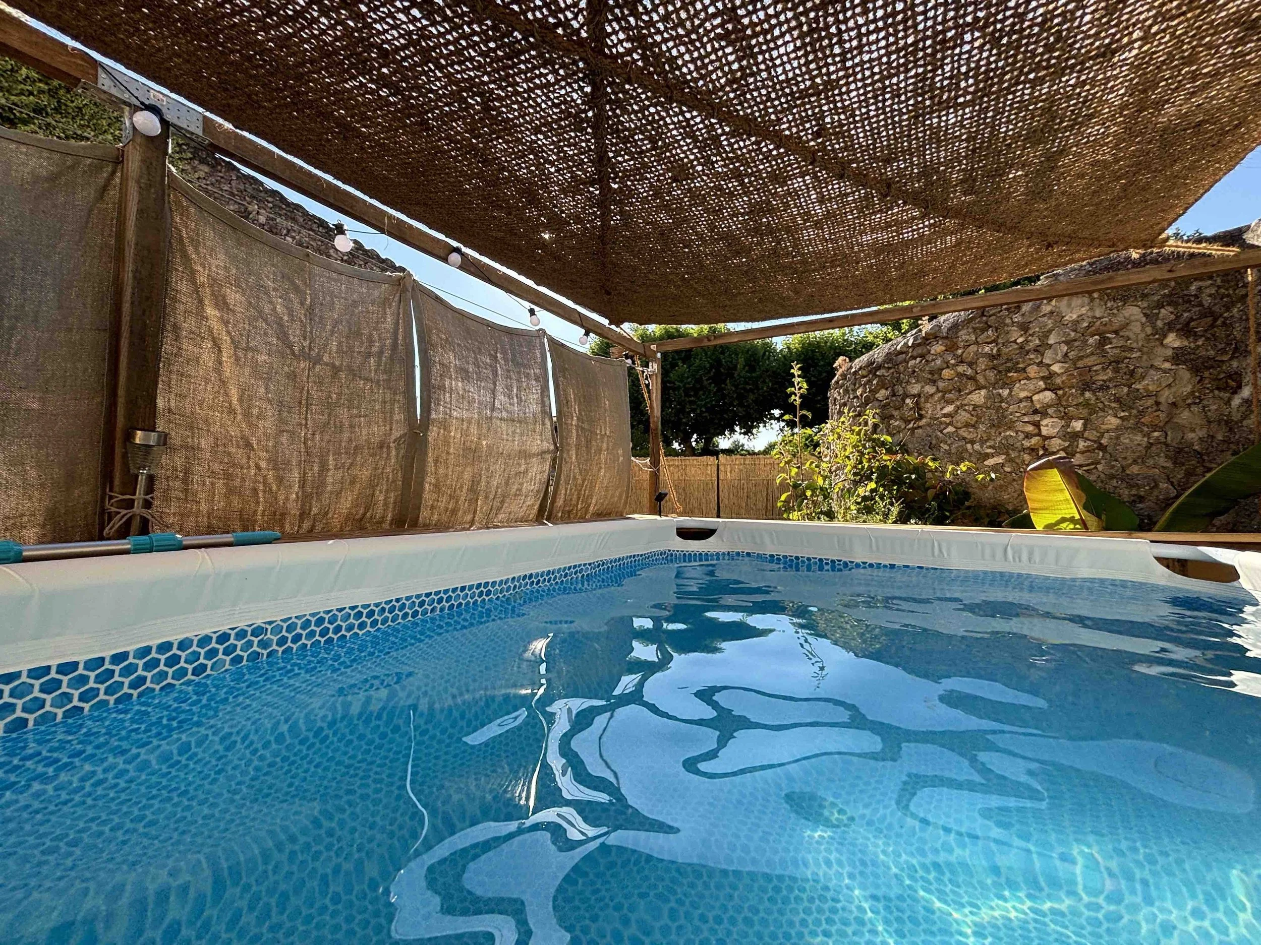 Une piscine avec un revêtement en mosaïque bleue, entourée d'une clôture en bambou et de murs en pierre, sous une pergola en bois avec une couverture en toile de jute.