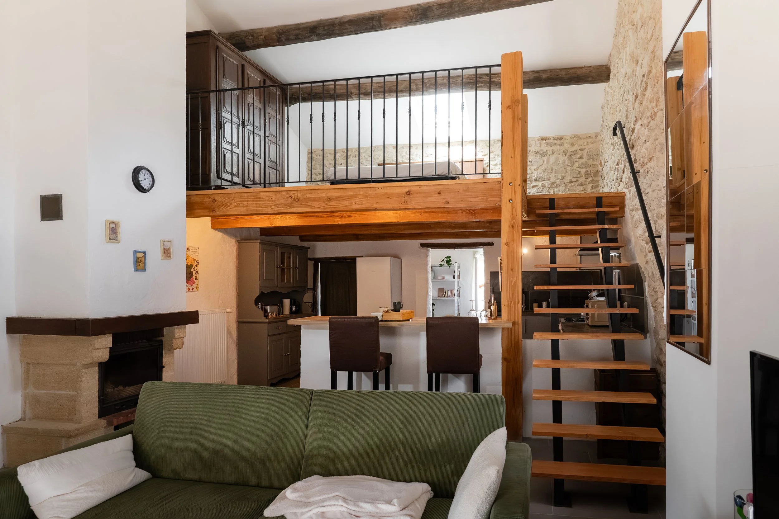 Intérieur d'un salon avec un canapé vert, une cheminée en pierre, une zone cuisine avec un plan de travail et des meubles gris, un escalier en bois et métal menant à une mezzanine avec un lit, murs en pierre et plafond avec poutres en bois.