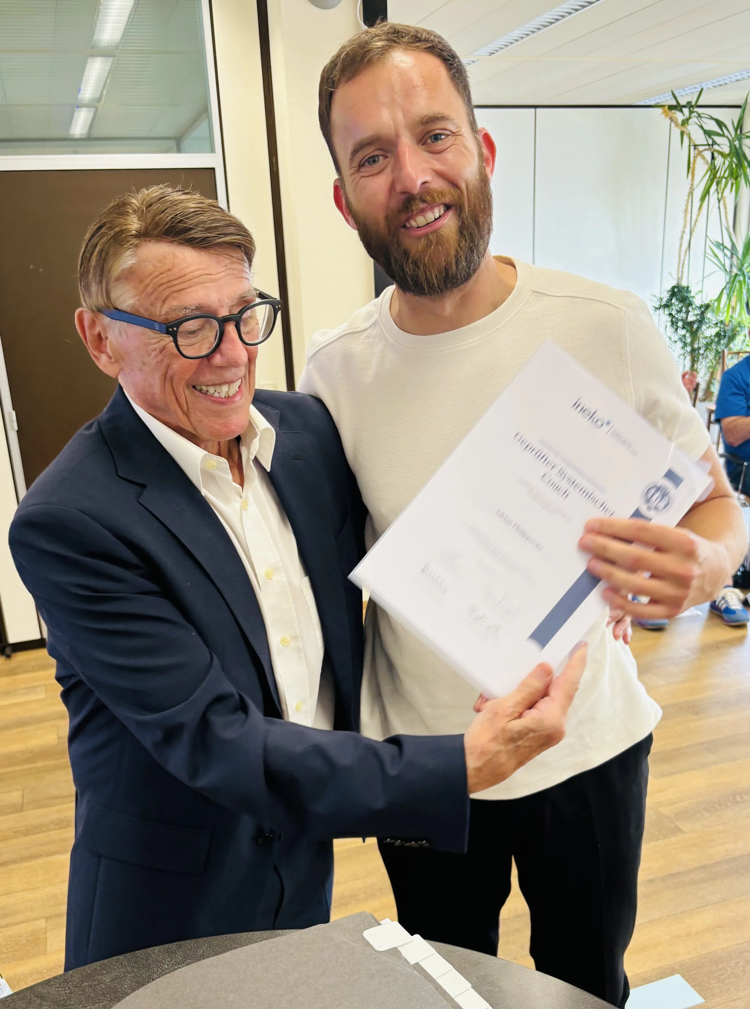 Zwei Männer bei einer Zertifikatsverleihung, der ältere Mann überreicht dem jüngeren einen Dokumentenordner, beide lächeln, im Hintergrund Pflanzen und andere Personen.