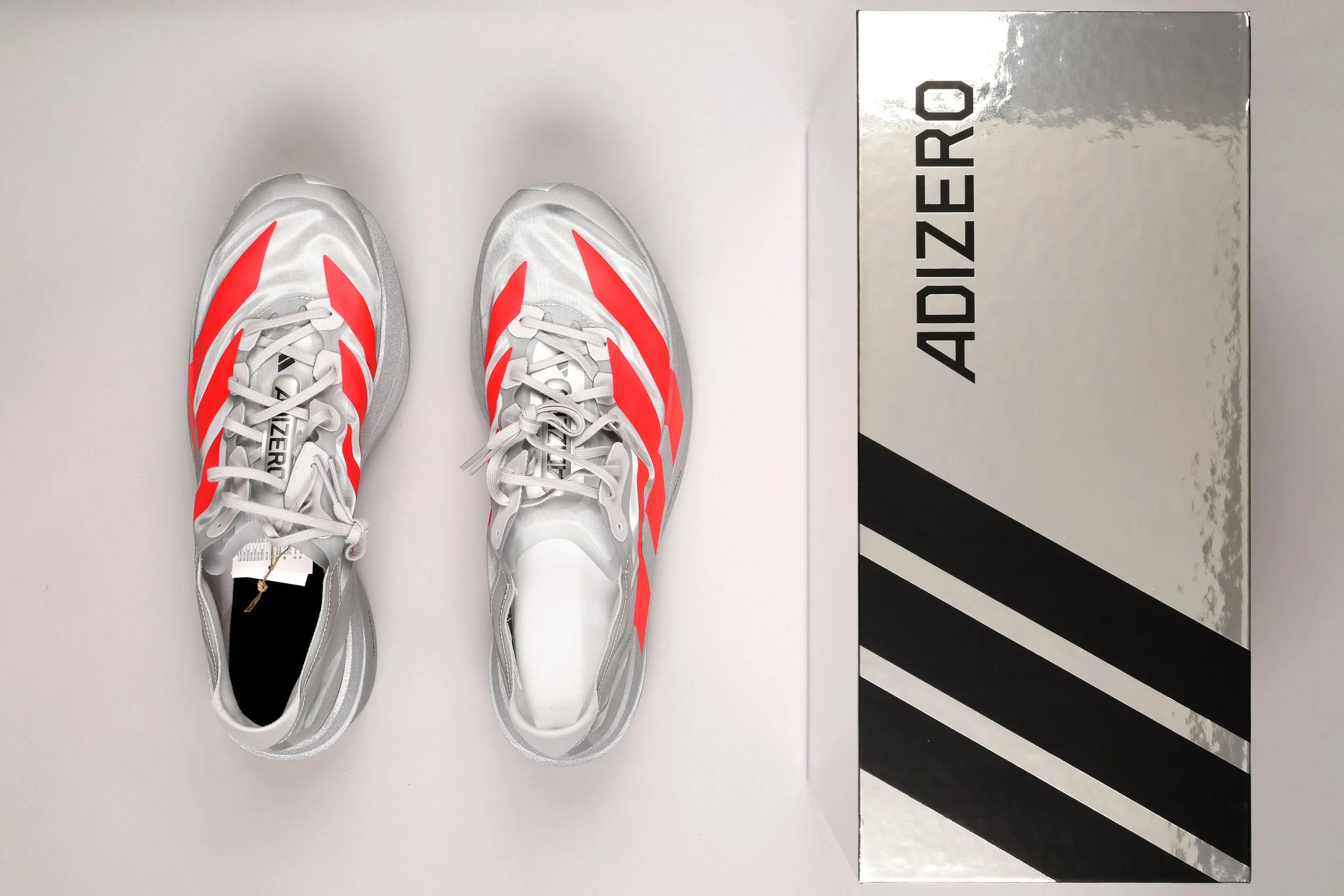 A pair of white Adidas Adizero running shoes with red stripes, placed side by side on a white surface next to a silver and black Adidas Adizero shoebox.