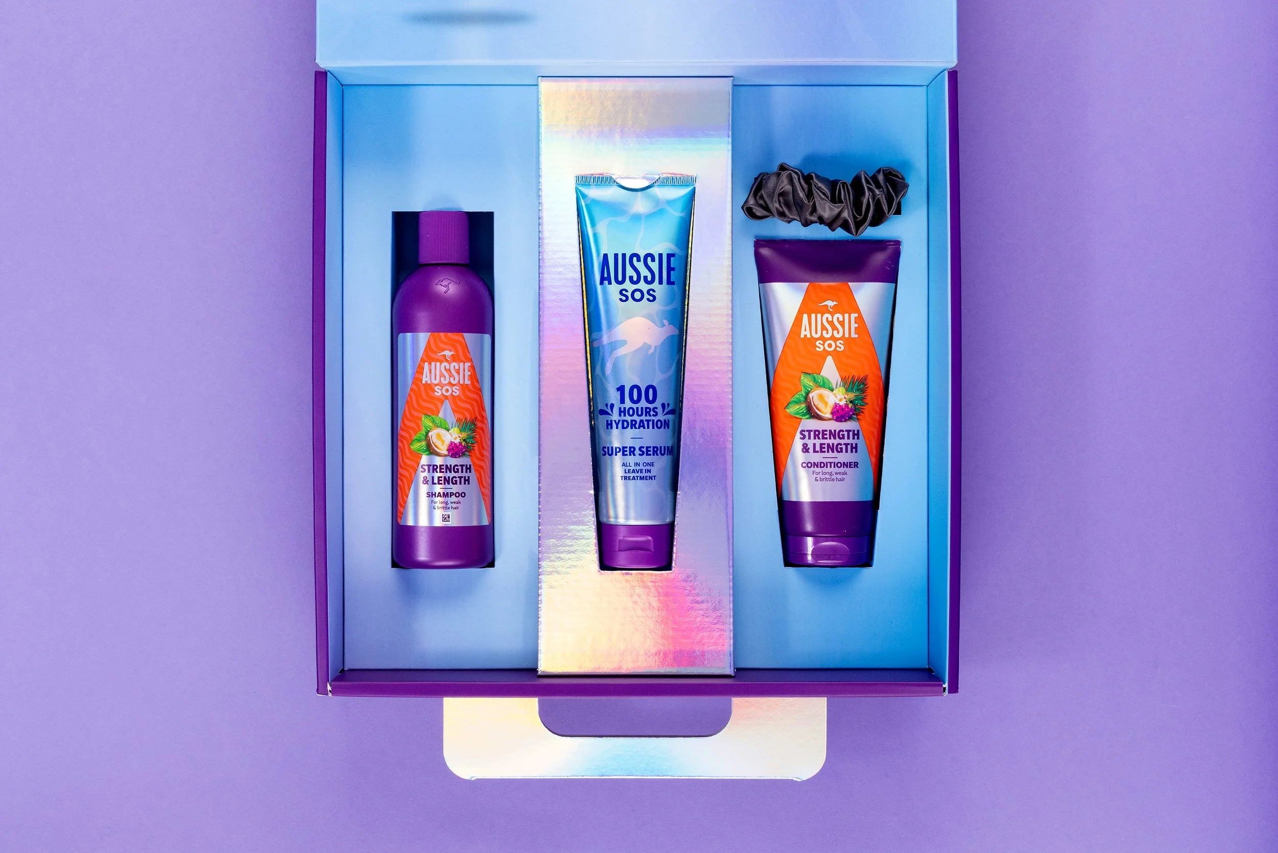 A set of Aussie hair care products, including shampoo, conditioner, and super serum, displayed in an iridescent box on a purple background.