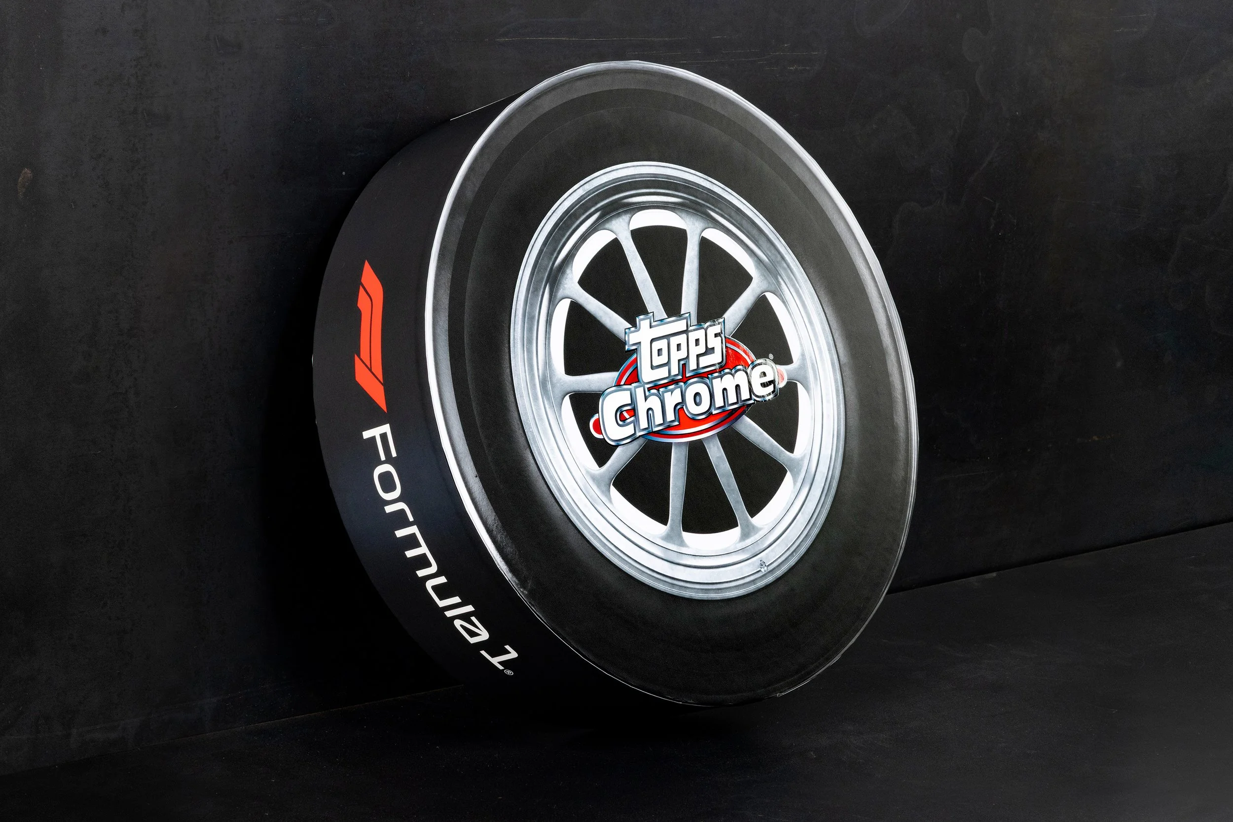 A racing tire with a silver alloy wheel and a black rubber tread. The tire features the brand 'Topps Chrome' in the center and the logo '4' along with the words 'F1 Definitive Collection' in white and red on the sidewall.