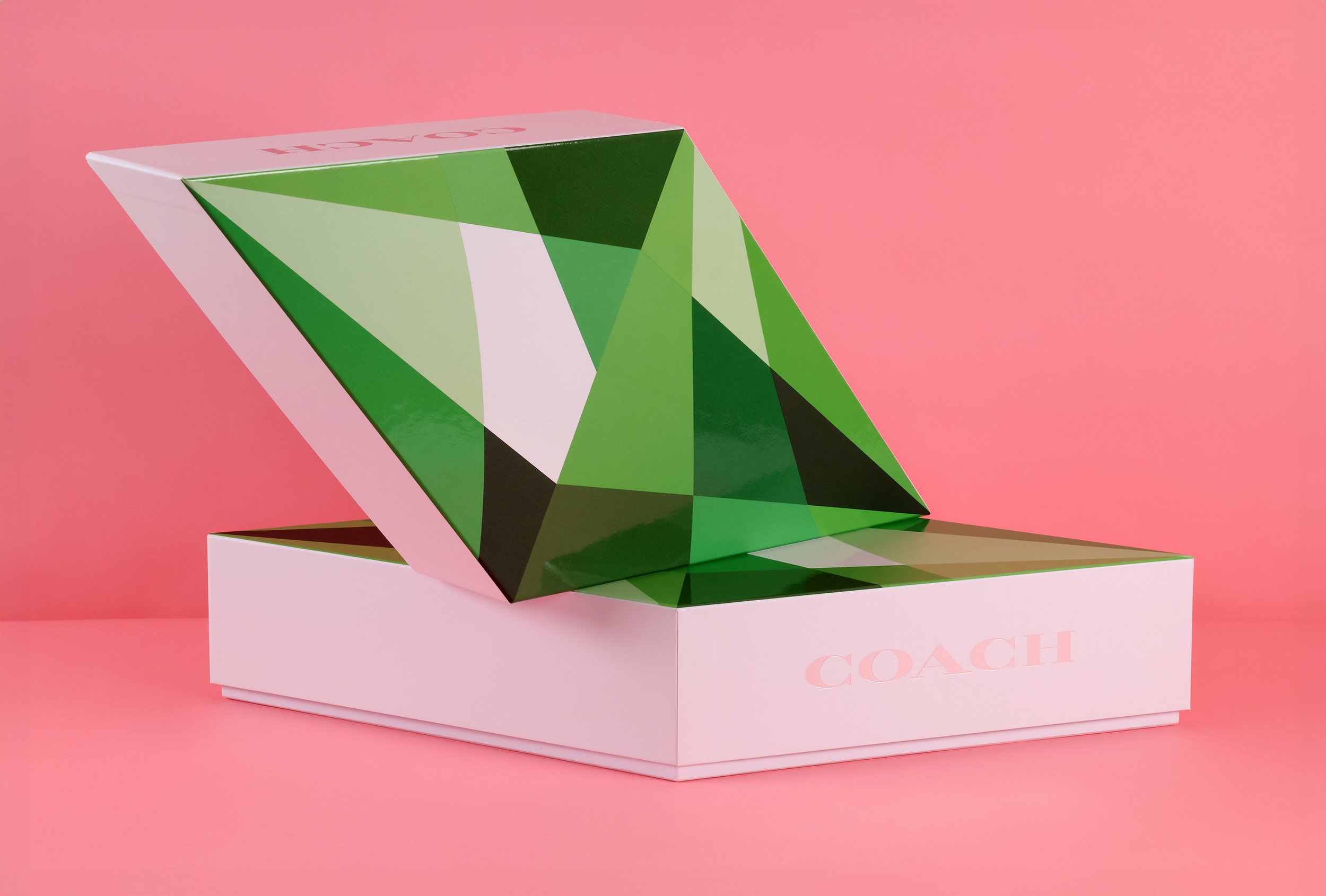 A geometric green and black jewelry box with a faceted surface, partially open on a pink background, with a pink and white COACH branded box underneath.