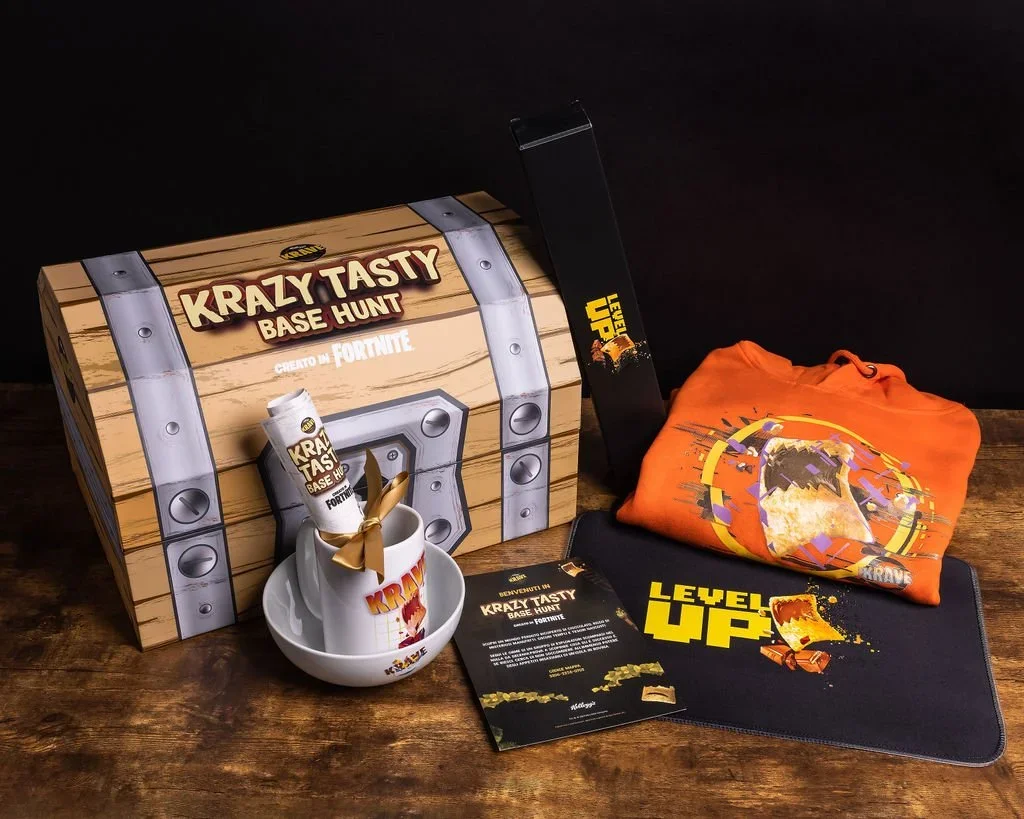 A Fortnite-themed loot box labeled 'Krazy Tasty Base Hunt' with Fortnite branding, a 'Level Up' cup, a rolled-up T-shirt, a black mat with 'Level Up' design, and a letter, all on a wooden surface.