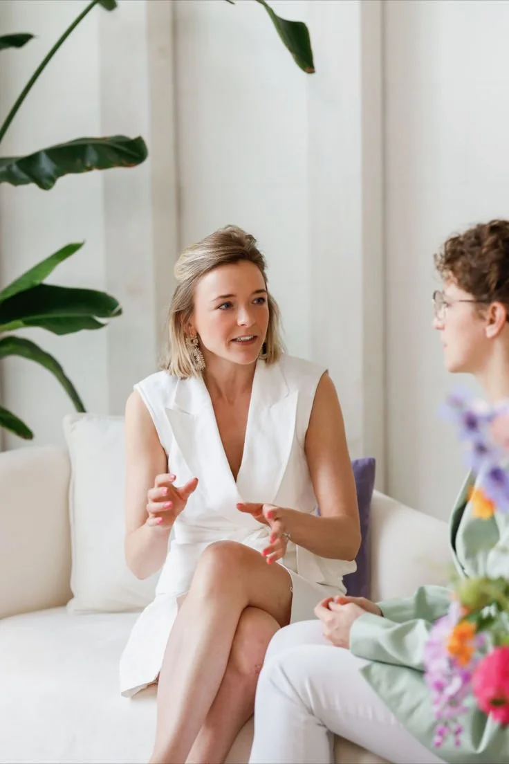 Twee vrouwen hebben een gesprek in een lichte, moderne kamer. Eén vrouw draagt een witte jurk en de ander draagt een groene jas en witte broek. Op de achtergrond grote groene planten.
