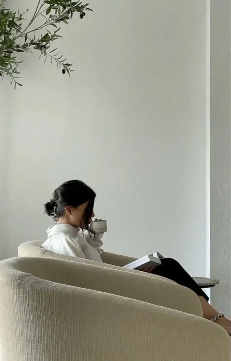 Vrouw die op een beige bank zit, een boek leest en een kopje vasthoudt, in een minimalistische kamer met een witte muur en een plant in de hoek.