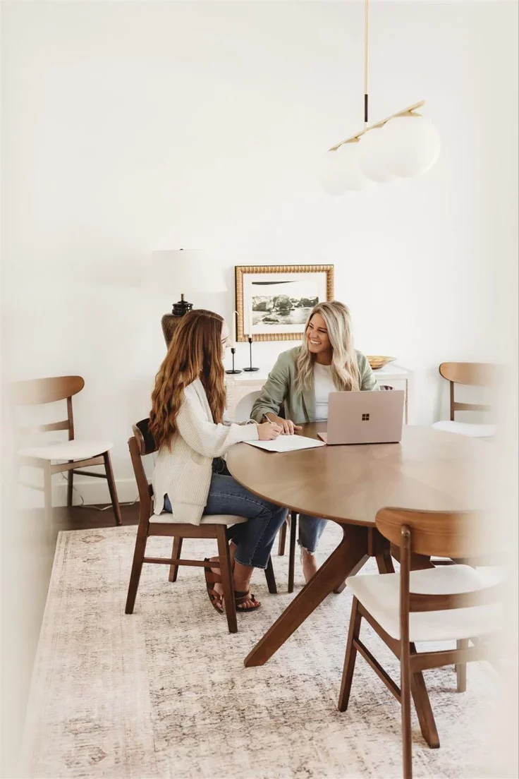 Twee vrouwen zitten aan een houten tafel in een lichte, stijlvolle kamer, praten en lachen. Er ligt een open laptop en een notitieboek op tafel.