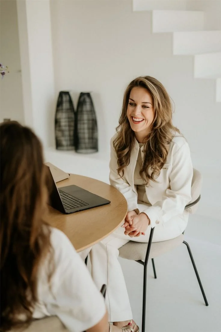Twee vrouwen die in gesprek zijn, zittend aan een ronde tafel met een laptop, in een lichte kamer