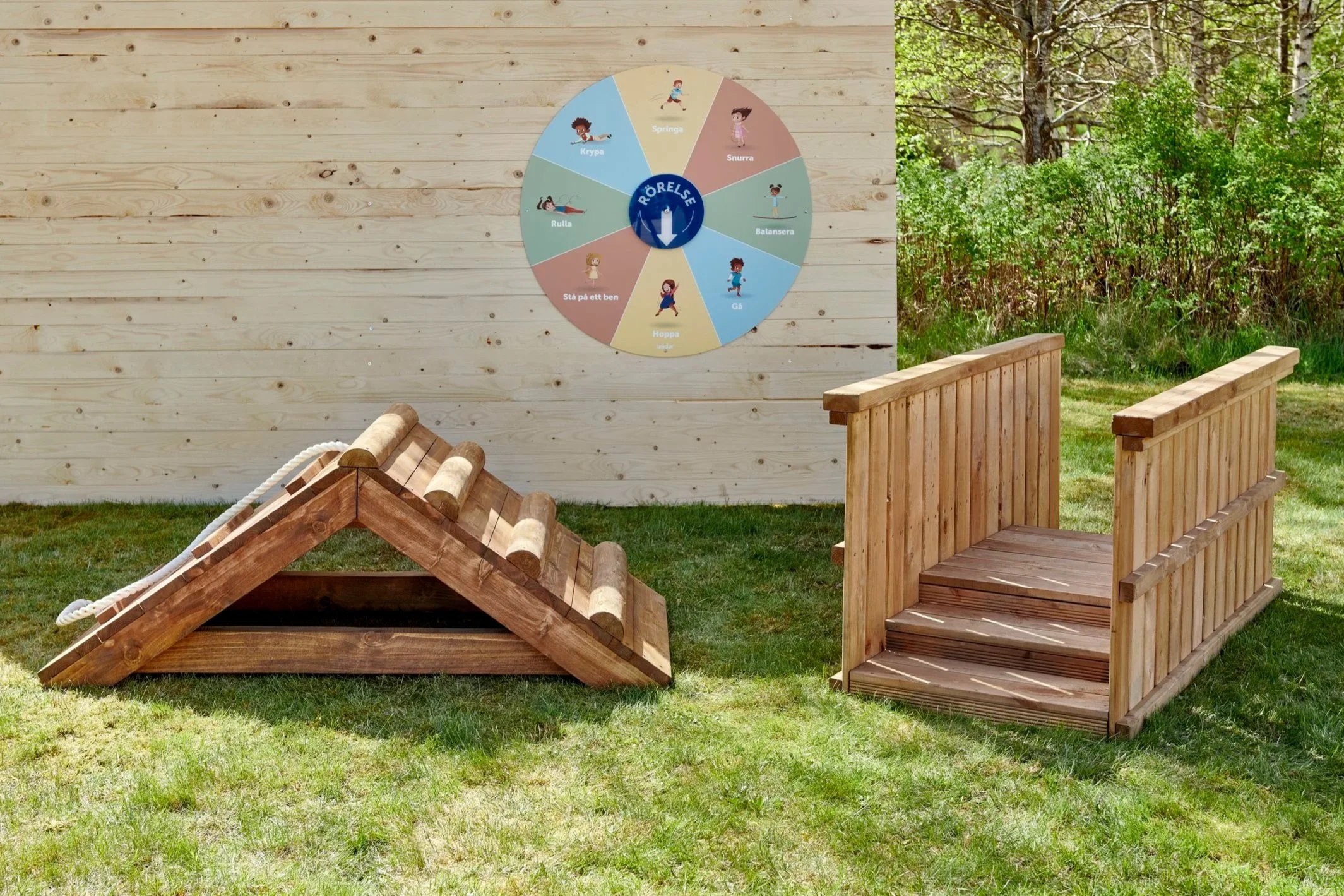 Et udendørs trælegeområde med en rund bevægelsescirkel på en træskive på en væg, der viser forskellige børneaktiviteter som springe, snurre, balancere, gå, hoppe, stå på ét ben, rulle og krybe. Der er også to træplatforme med trapper. Omgivelserne består af grønne græsplæner og træer.