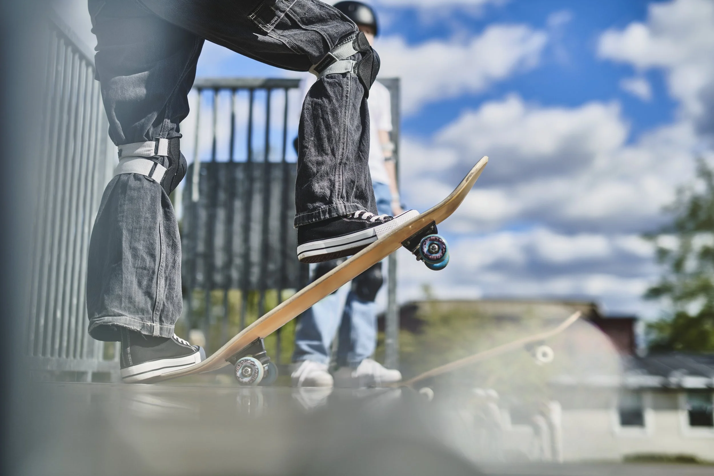 En person på skateboard står på en rampe, med en anden person i baggrunden. Begge personer er iført jeans og sneakers, og den ene person holder en skateboard.