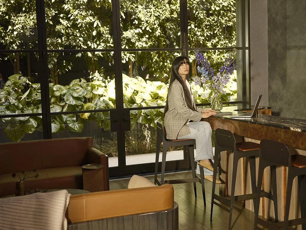 A woman with long dark hair and light-colored pants sits at a high counter with a laptop, gazing outside at a lush green garden through large glass windows. There are flowers in a vase on the counter, and a cozy seating area with a brown leather couch and pillows in the foreground.