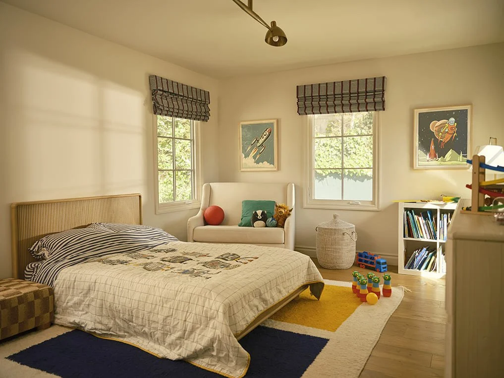 Child's bedroom with a bed, two windows, artwork, toys, and a bookshelf.