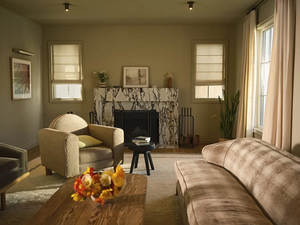 Living room with beige armchair and sofa, marble fireplace, wooden coffee table with flowers, windows with beige curtains, potted plant, framed wall art, and ceiling lights.
