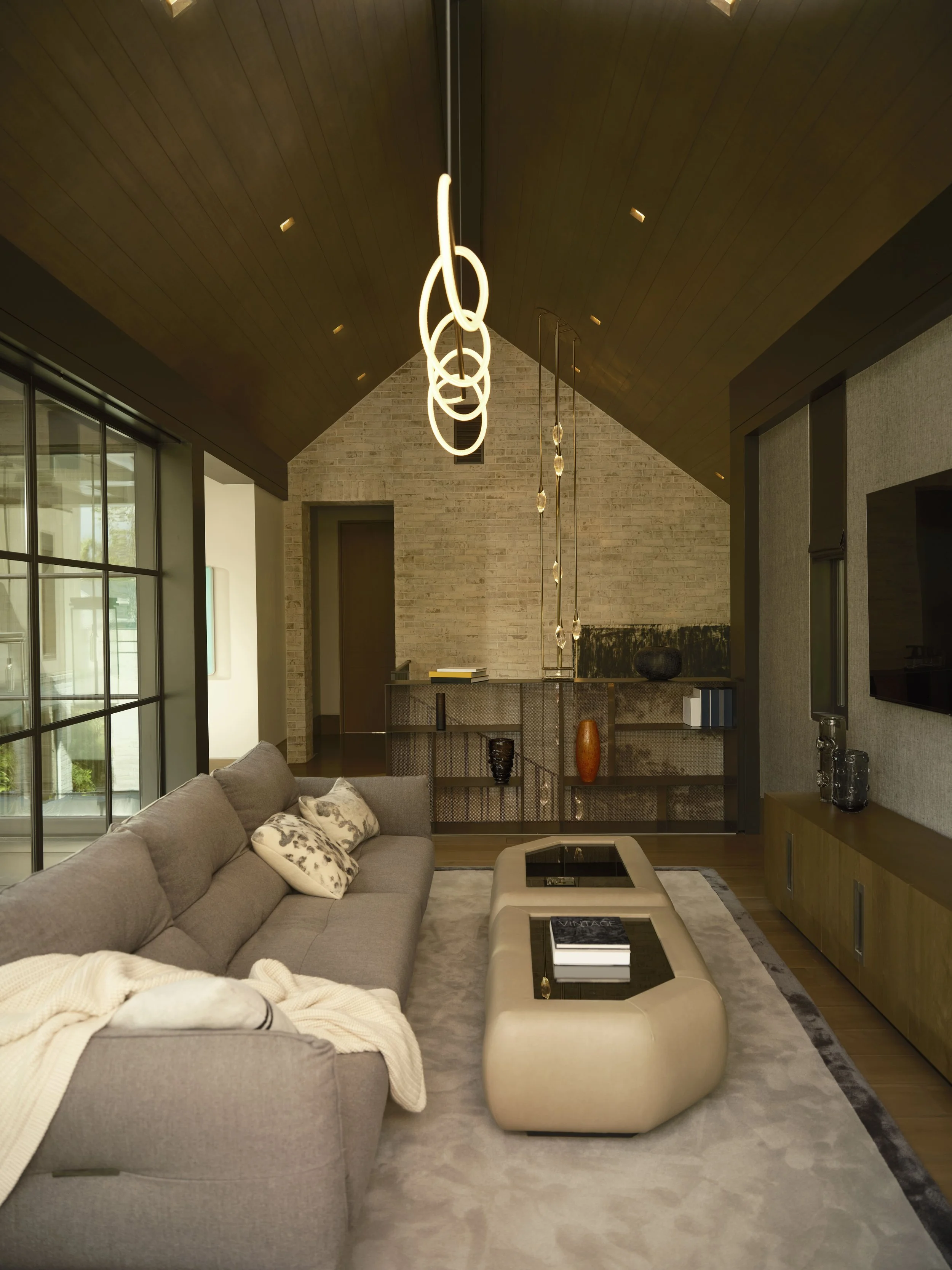 Modern living room with a gray sofa, decorative pillows, a cream-colored coffee table with books, a textured area rug, wood flooring, large windows, black lighting fixtures, and a brick wall in the background.