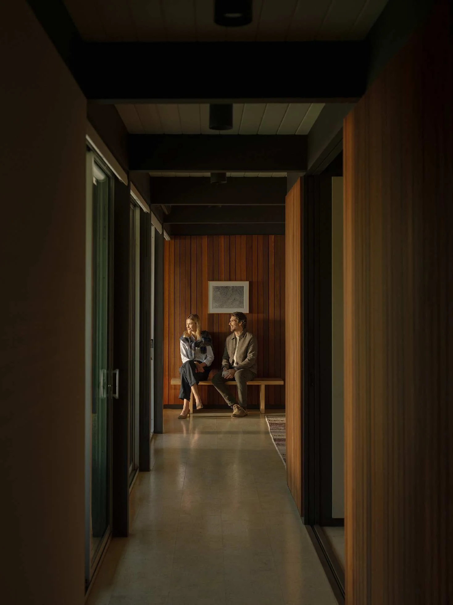 Interior view of a modern building corridor with wood-paneled walls, a bench, and two people sitting and conversing, dressed in business attire.
