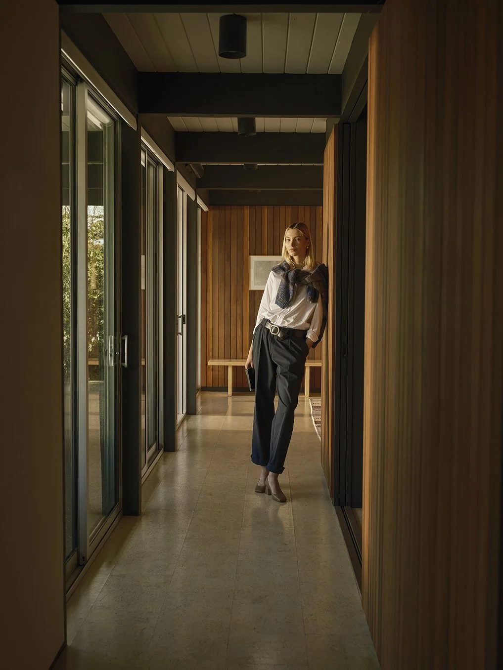 A woman stands in a hallway with wooden wall paneling, wearing a white blouse, black trousers, beige high heels, and a dark sweater draped over her shoulders, holding a book.