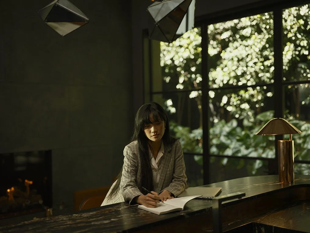 A woman in a checkered blazer sits at a dark wooden desk in a dimly lit room, writing in a notebook, with a large window behind her showing lush green trees outside, and a brass table lamp on the right side.