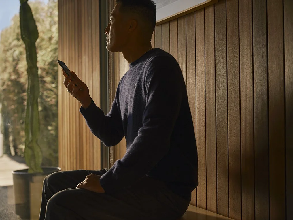 A man sitting indoors by a wooden wall, looking at his phone, with a large window and a cactus plant in the background.