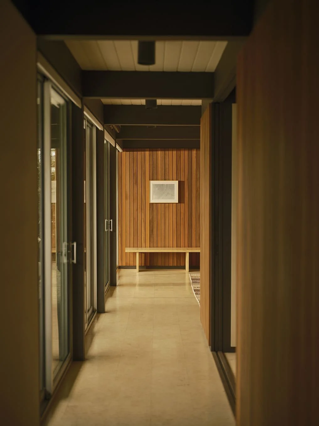 Indoor corridor with wooden walls and sliding glass doors on the left, a bench at the end of the corridor, and a framed picture on the wood-paneled back wall.