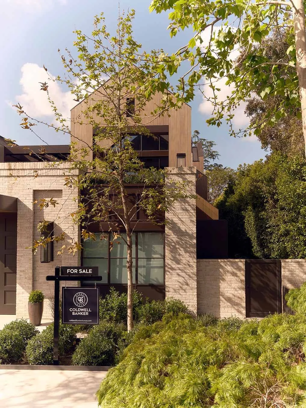 Modern multi-story house with a "For Sale" sign in front, surrounded by greenery and trees, under a partly cloudy sky.