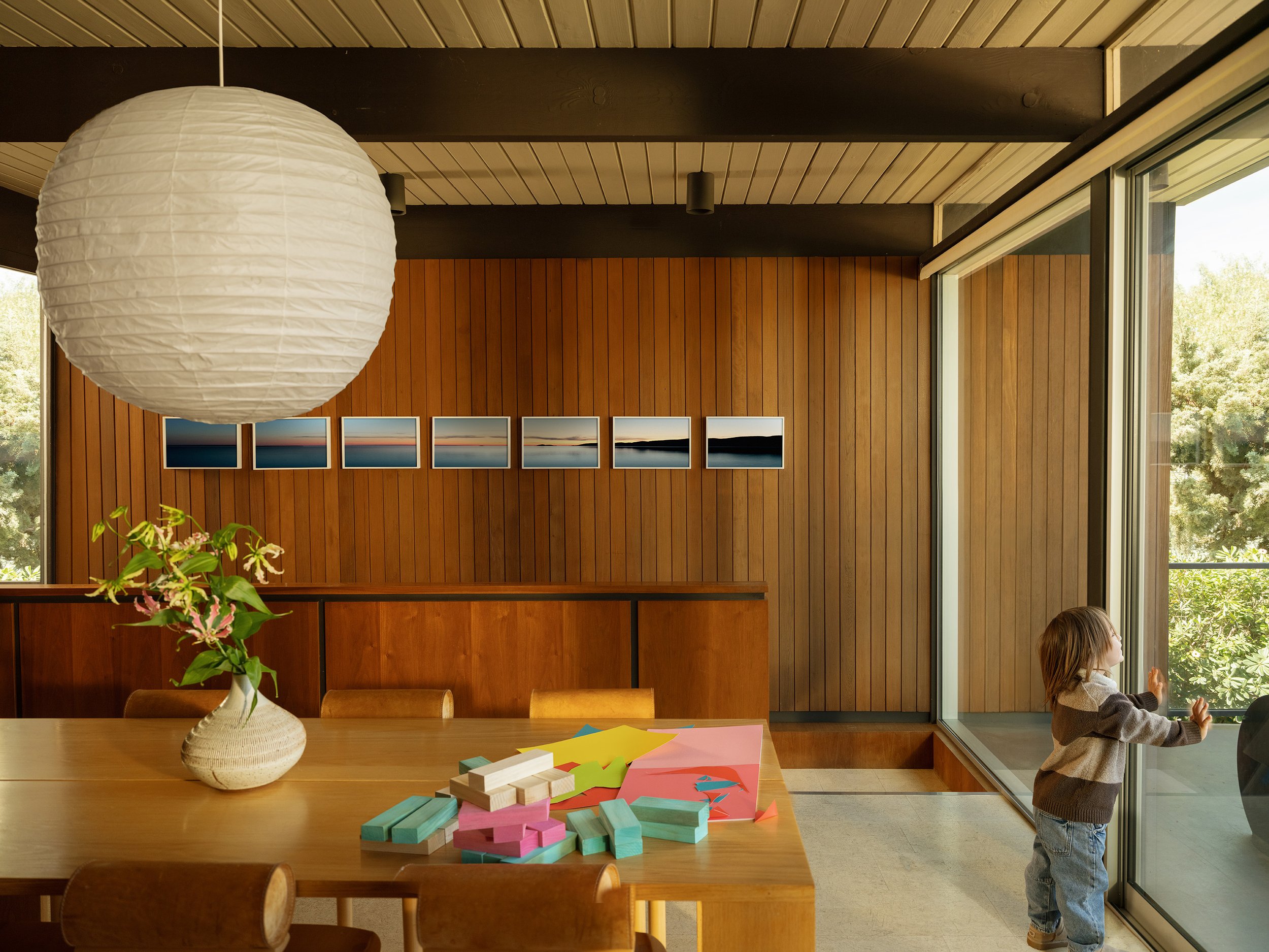 A dining area with a large white paper lantern hanging from the ceiling, a wooden table with colorful wooden blocks and papers, a vase with pink flowers, and a child playing by a large window showing green trees outside.