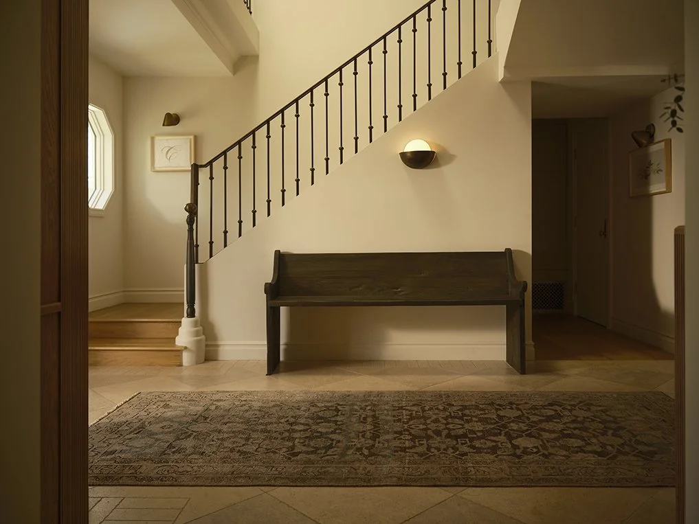 Interior view of a staircase landing area with a wooden bench, wall-mounted light fixture, and framed artwork, featuring a small window to the left.