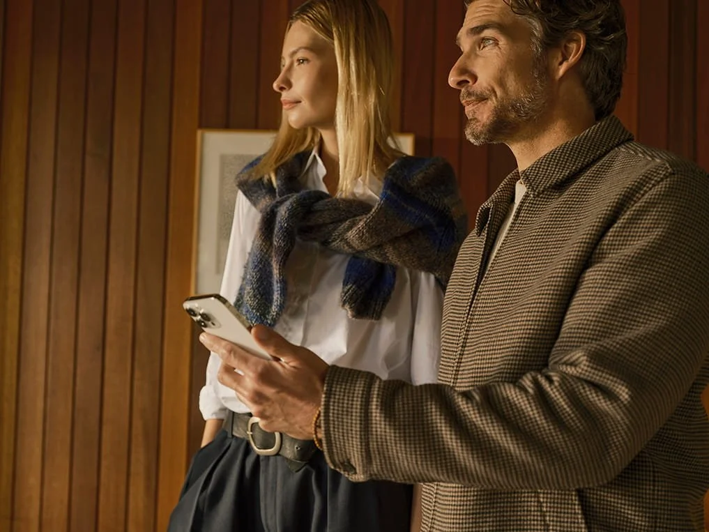A man and a woman standing indoors against a wooden wall, with the man holding a smartphone, both looking to their right.