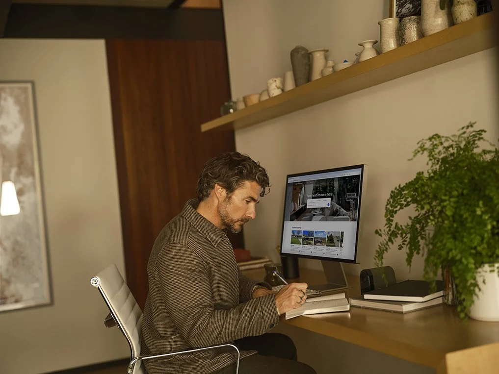 Man working at a home office desk with a computer, notebooks, and a potted plant.