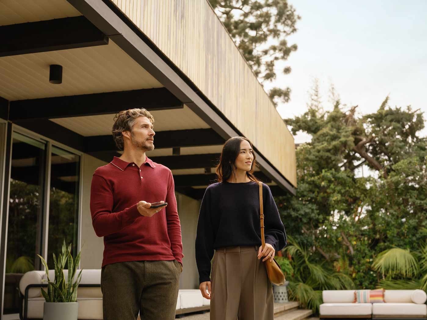 A man and woman standing outdoors near a modern house, looking to the side, with trees and greenery in the background.
