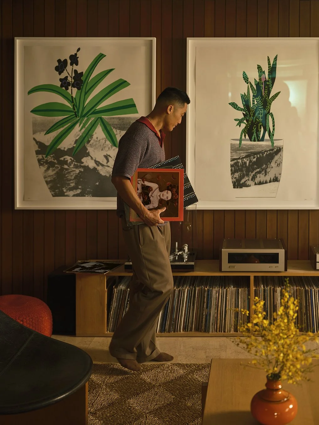 A man in a gray shirt and khaki pants is holding vinyl records in a room with wooden walls, standing in front of two large framed botanical illustrations and a shelf full of vinyl records.