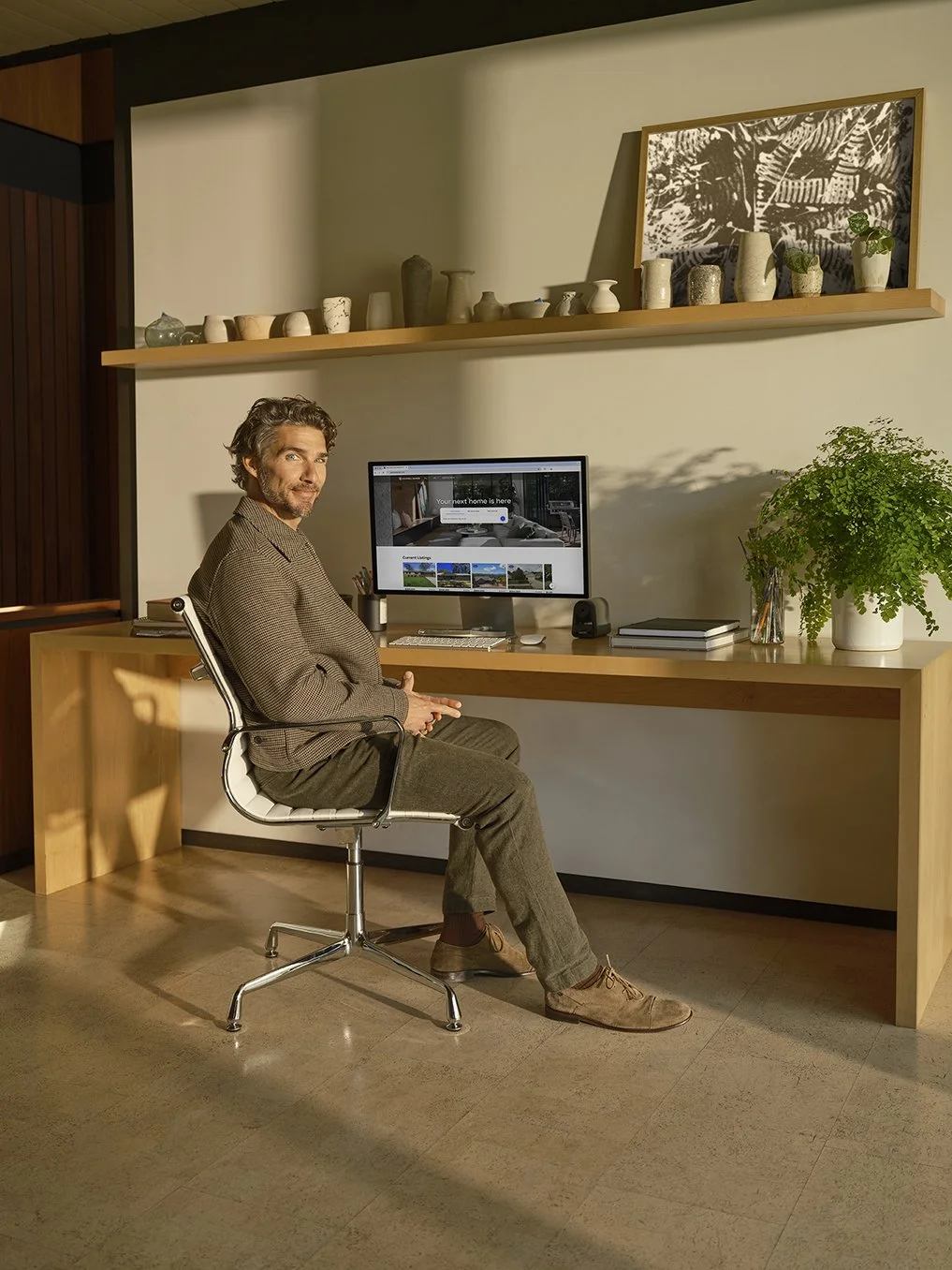 A man with gray hair and a beard sits at a wooden desk with a computer monitor, a potted plant, and some books. There are shelves with vases and a framed picture on the wall behind him, in a modern office or home workspace.