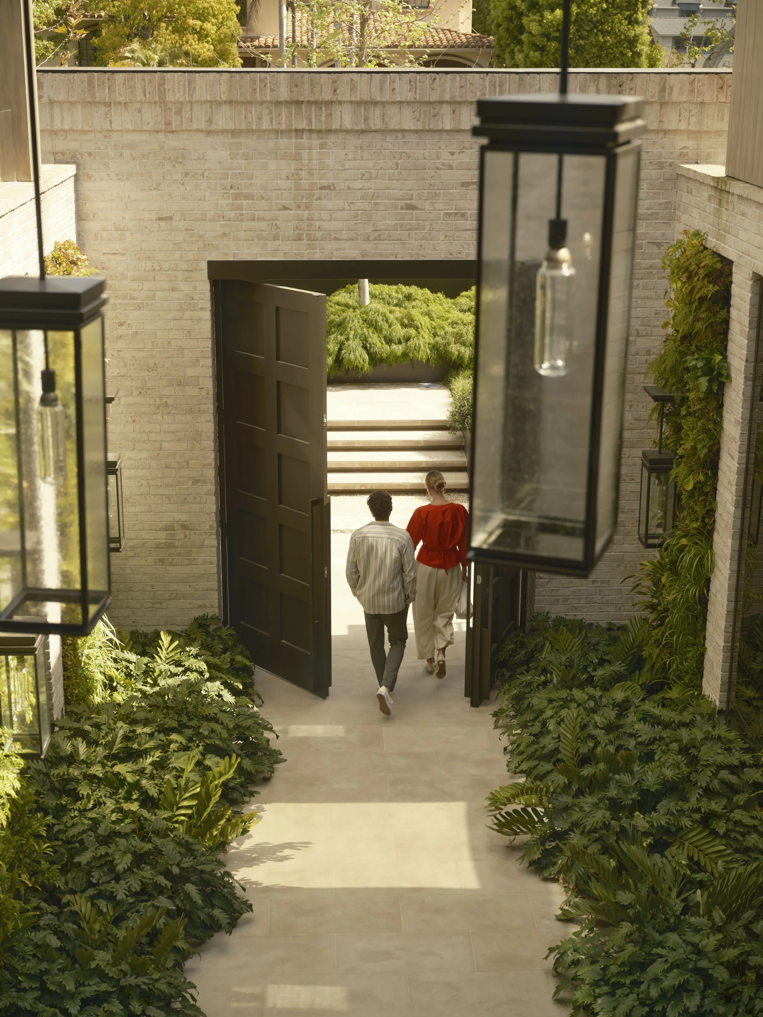 Two people walking out of a modern house with greenery surrounding the entrance, viewed through an open black door with glass lantern lights hanging on each side.