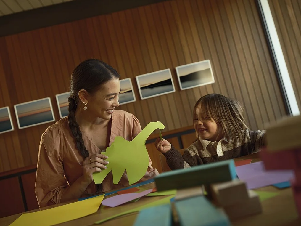 A woman and a young girl sitting at a table with colorful paper and books, smiling and playing with a green paper dinosaur in a room with wood-paneled walls and framed landscape photos.