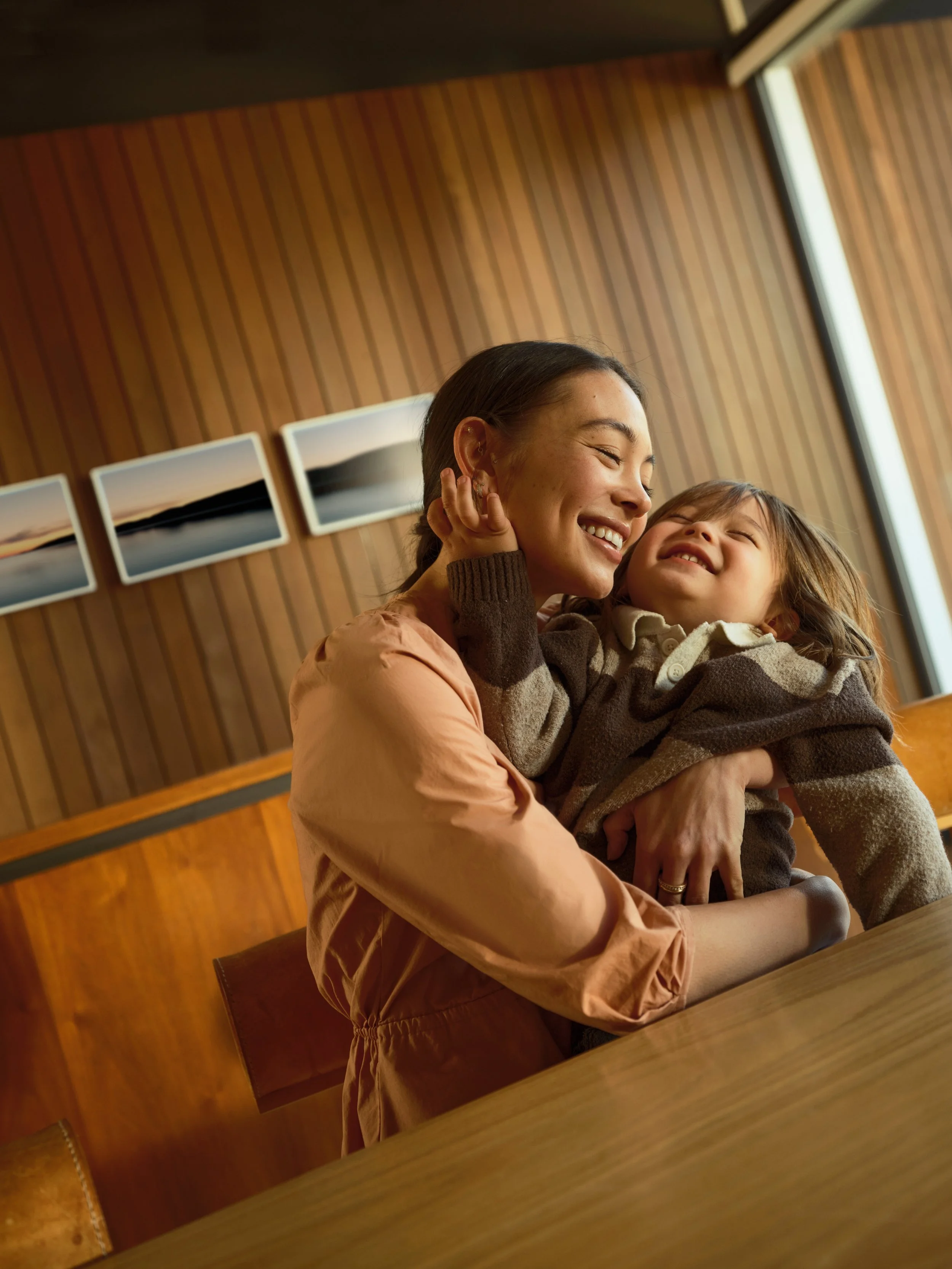 A woman and a young girl smiling and hugging in a cozy wooden room with framed pictures on the wall.