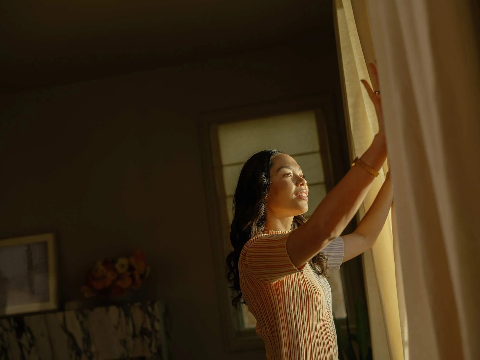 A woman with long dark hair and a striped shirt is reaching out and touching a wall or window, sunlight illuminating her face in a cozy indoor setting.